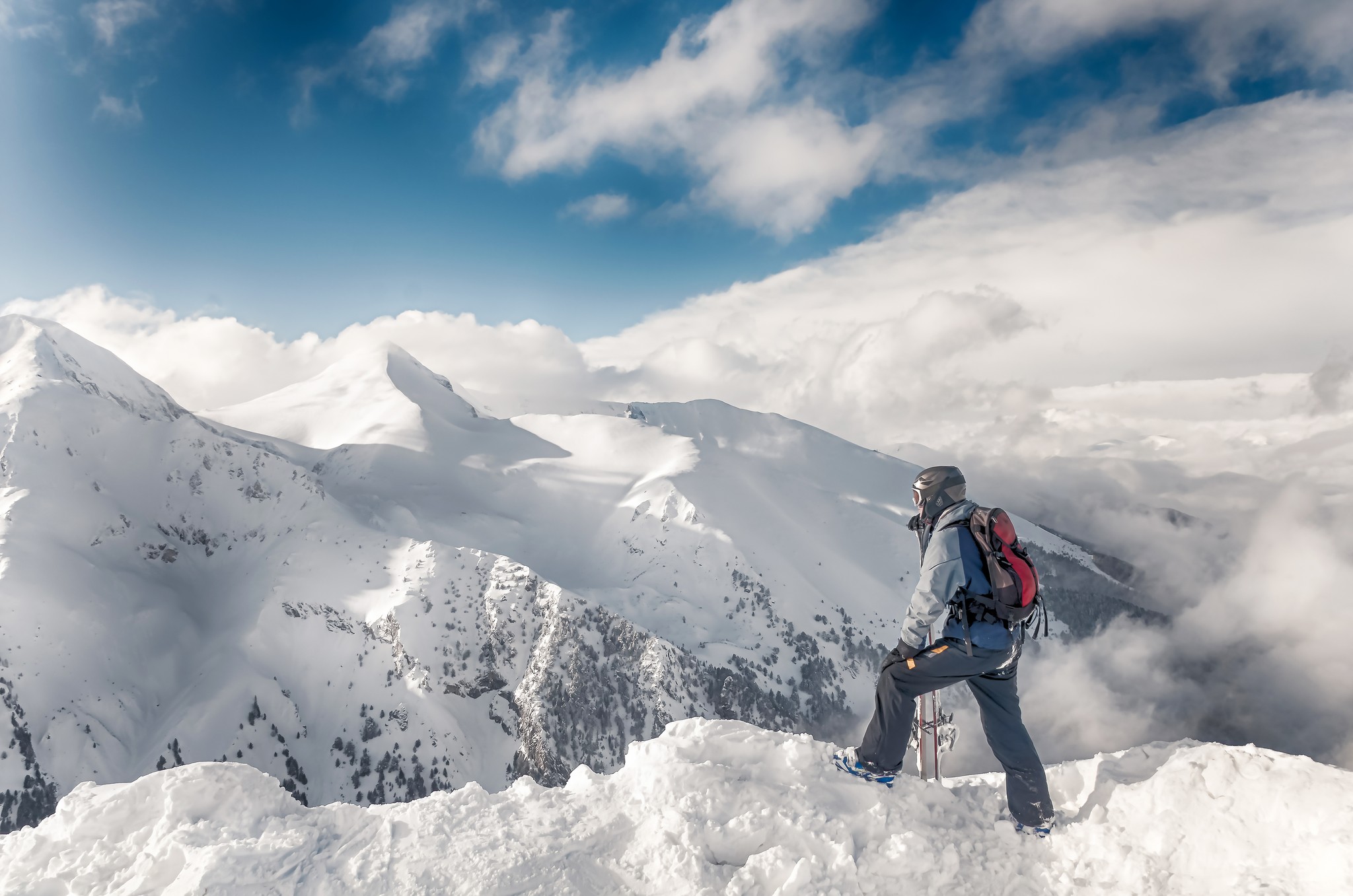 A skier looks out at a panoramic view of a European mountain range covered in fresh snow under a clear winter sky.