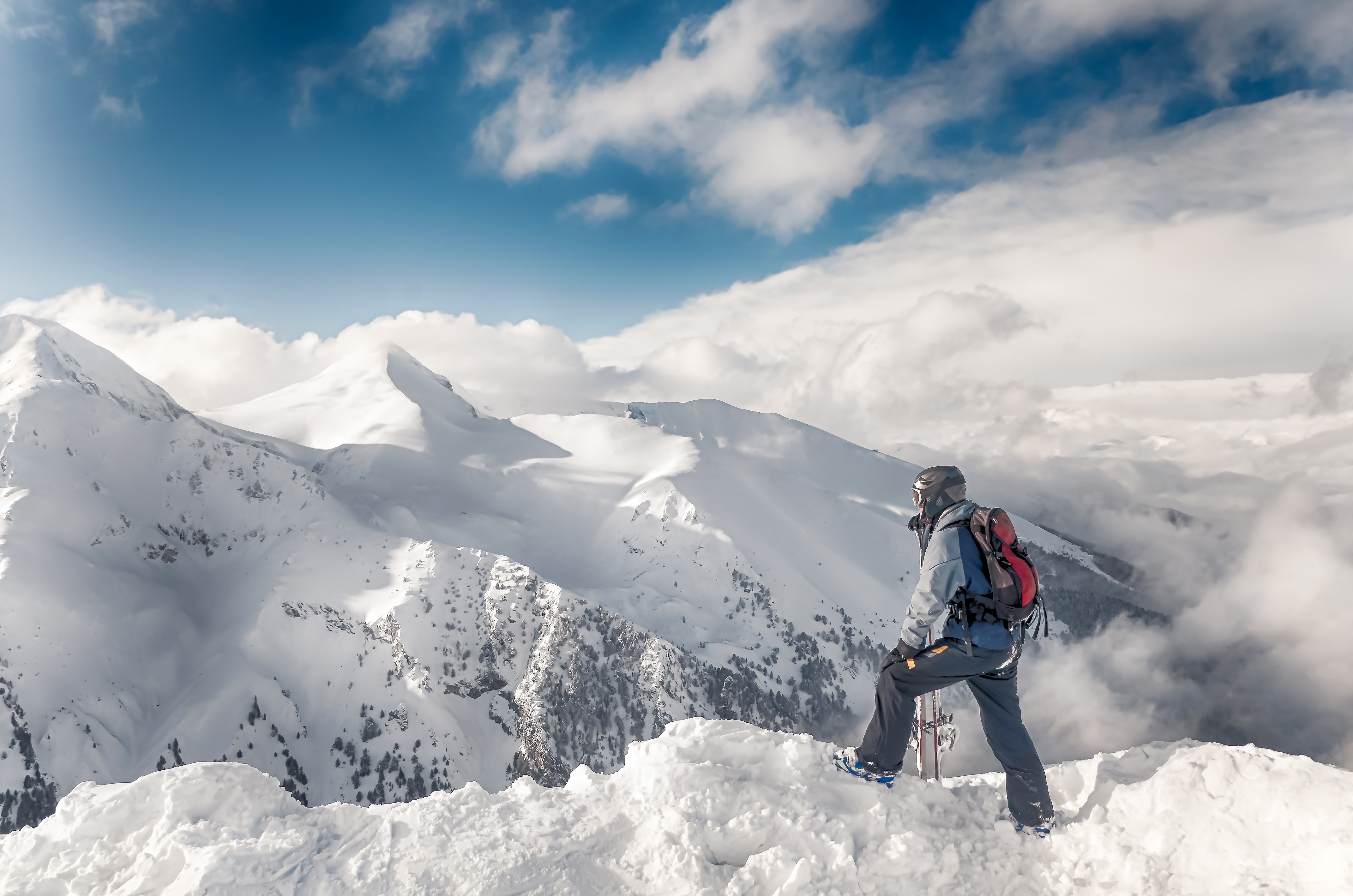 A skier looks out at a panoramic view of a European mountain range covered in fresh snow under a clear winter sky.