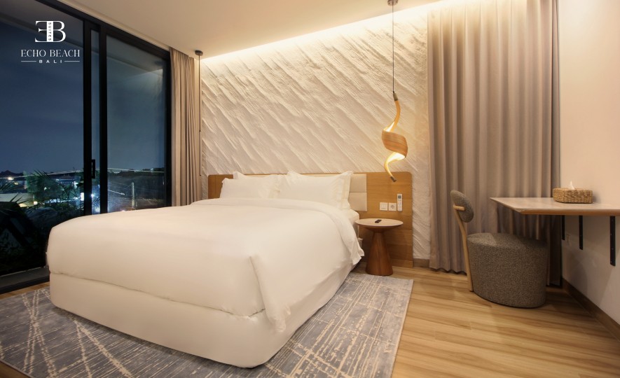 Echo Beach Suite Bedroom at Night: Cozy nighttime view of the white bed, backlit textured wall, and unique hanging light fixture, visible through the large window. Luxury accommodation in Canggu, Bali.