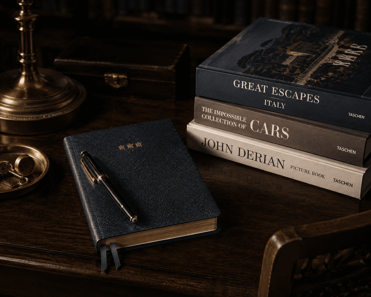 A dark wood desk scene featuring a textured navy blue leather-bound journal embossed with three small gold crowns, paired with a black and gold fountain pen. To the right, a stack of three Taschen books includes "Great Escapes Italy," "The Impossible Collection of Cars," and "John Derian Picture Book." A brass lamp base, a small wooden box, and a brass tray with vintage keys complete the moody composition.