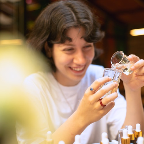 Isang bisita na nakangiti habang maingat na nagbubuhos ng mga sangkap ng pabango sa maliit na basong panukat sa panahon ng isang workshop.