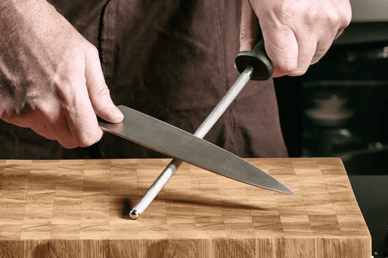 A person honing a kitchen knife with a steel honing rod, resting the tip on an end grain cutting board for added stability