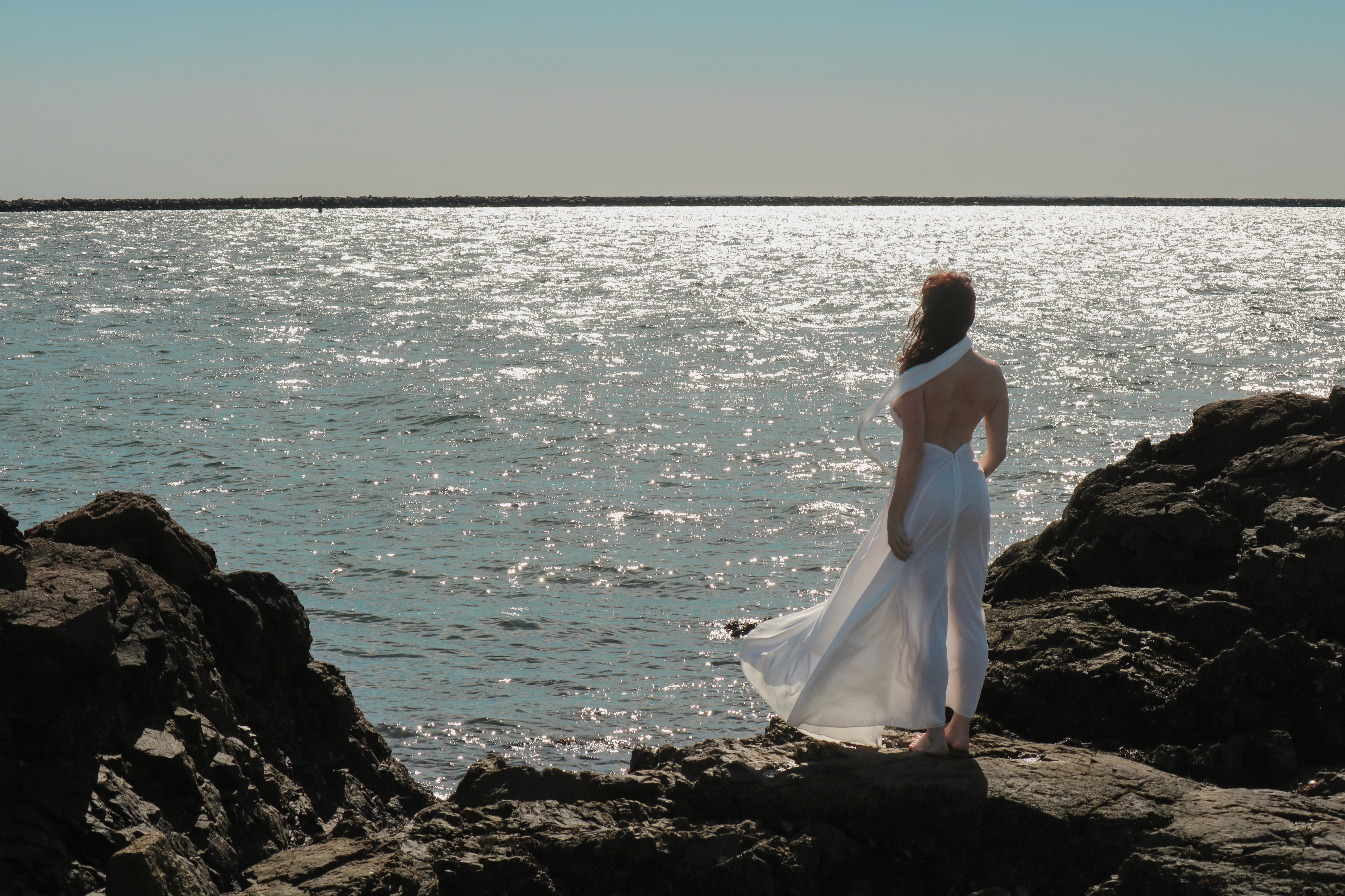 Hyperborea — a model in white standing on rocks above the sea.