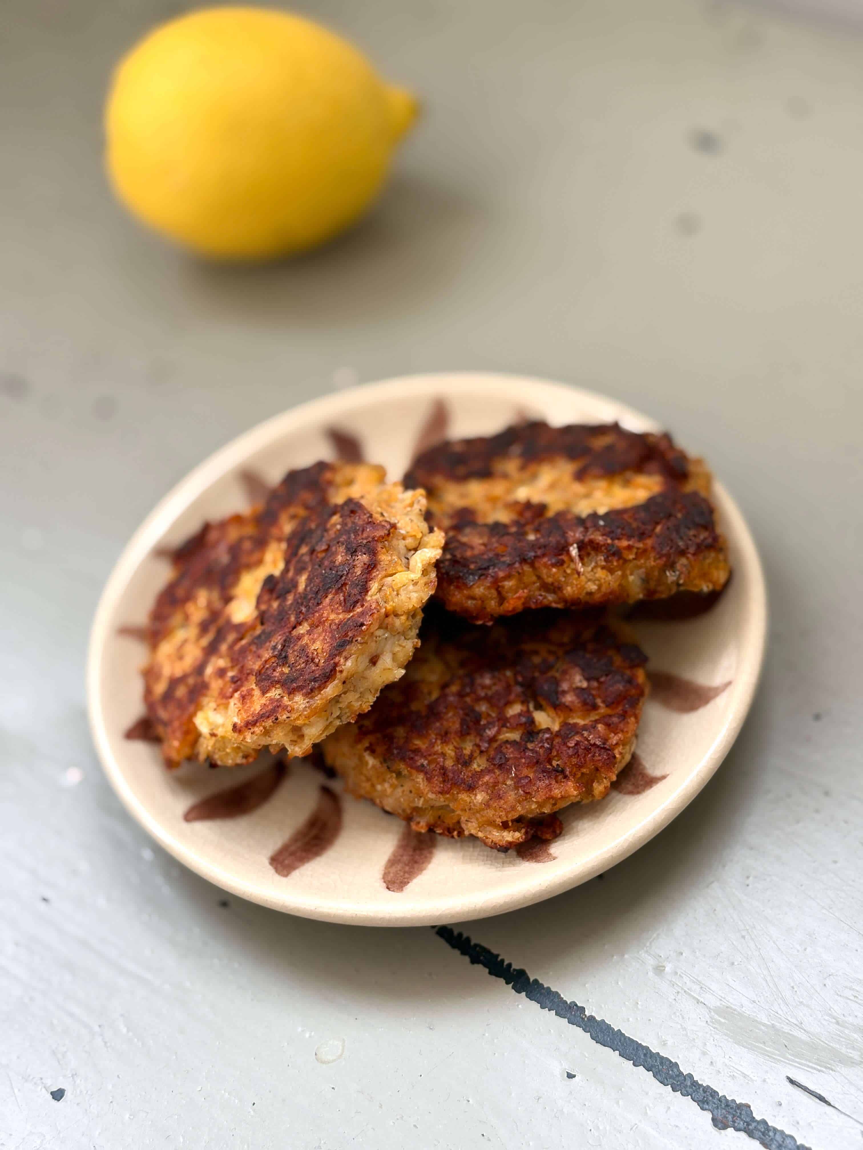 Golden-brown sardine fritters stacked on a small ceramic plate, with a lemon in the background.