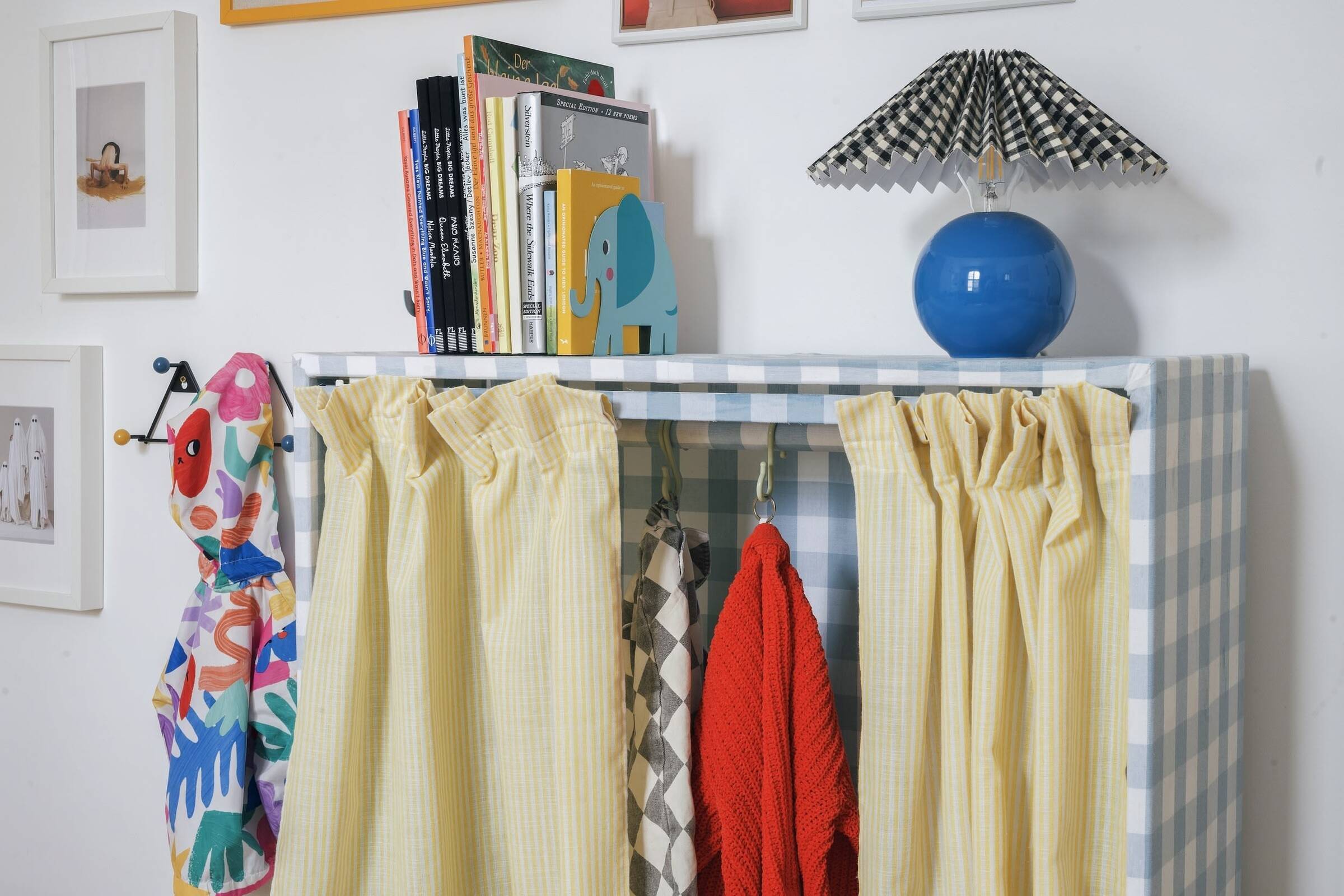 Contrasting patterns working together: an accent table with yellow and white striped curtains, colorful towels hanging, a bookshelf with children books, a blue round lamp with a black and white checkered lampshade, and framed artwork on the wall