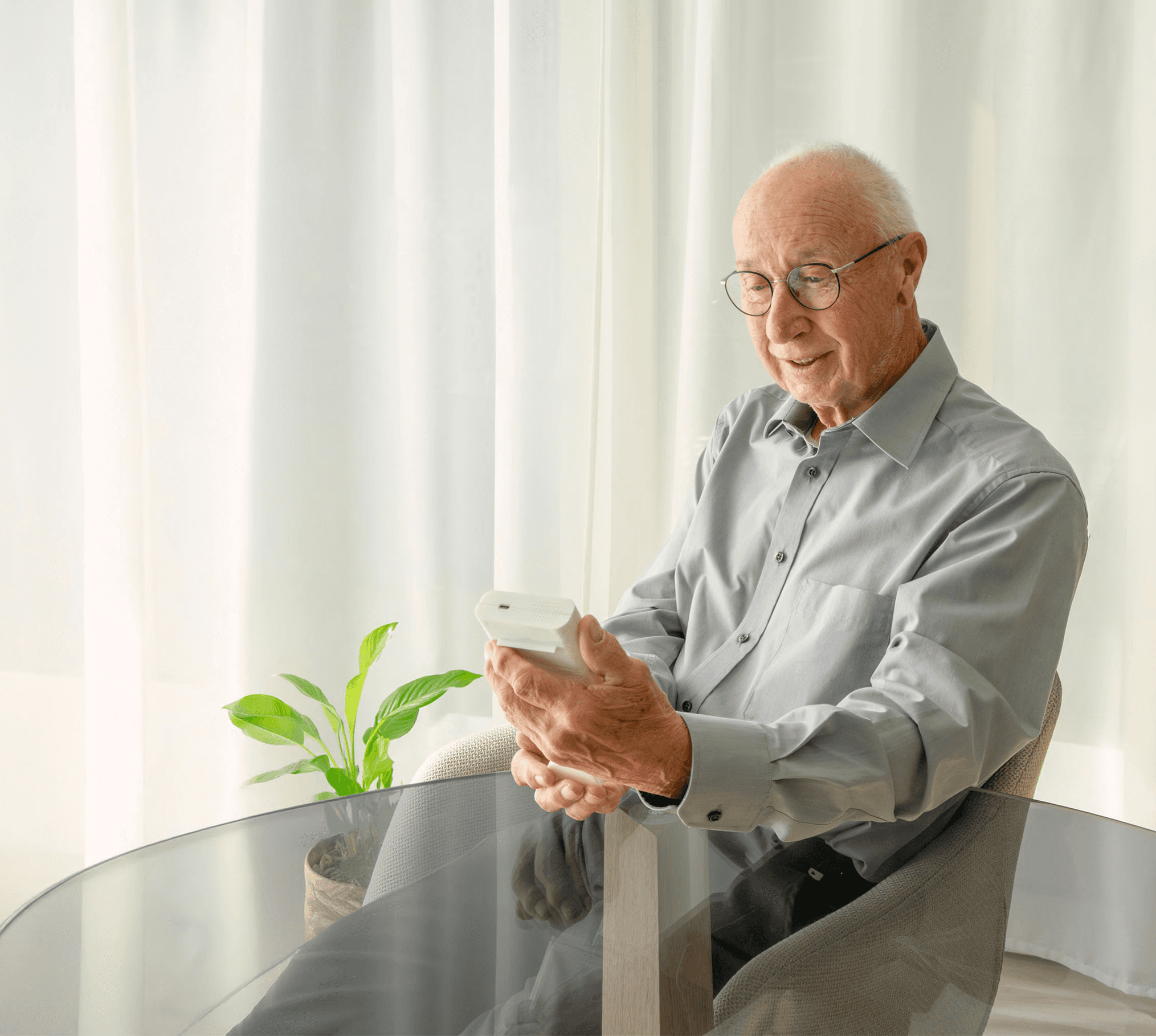 A smiling elderly man, wearing glasses, sitting at a table and holding a white smart medication dispenser (Medicase) in his hands.