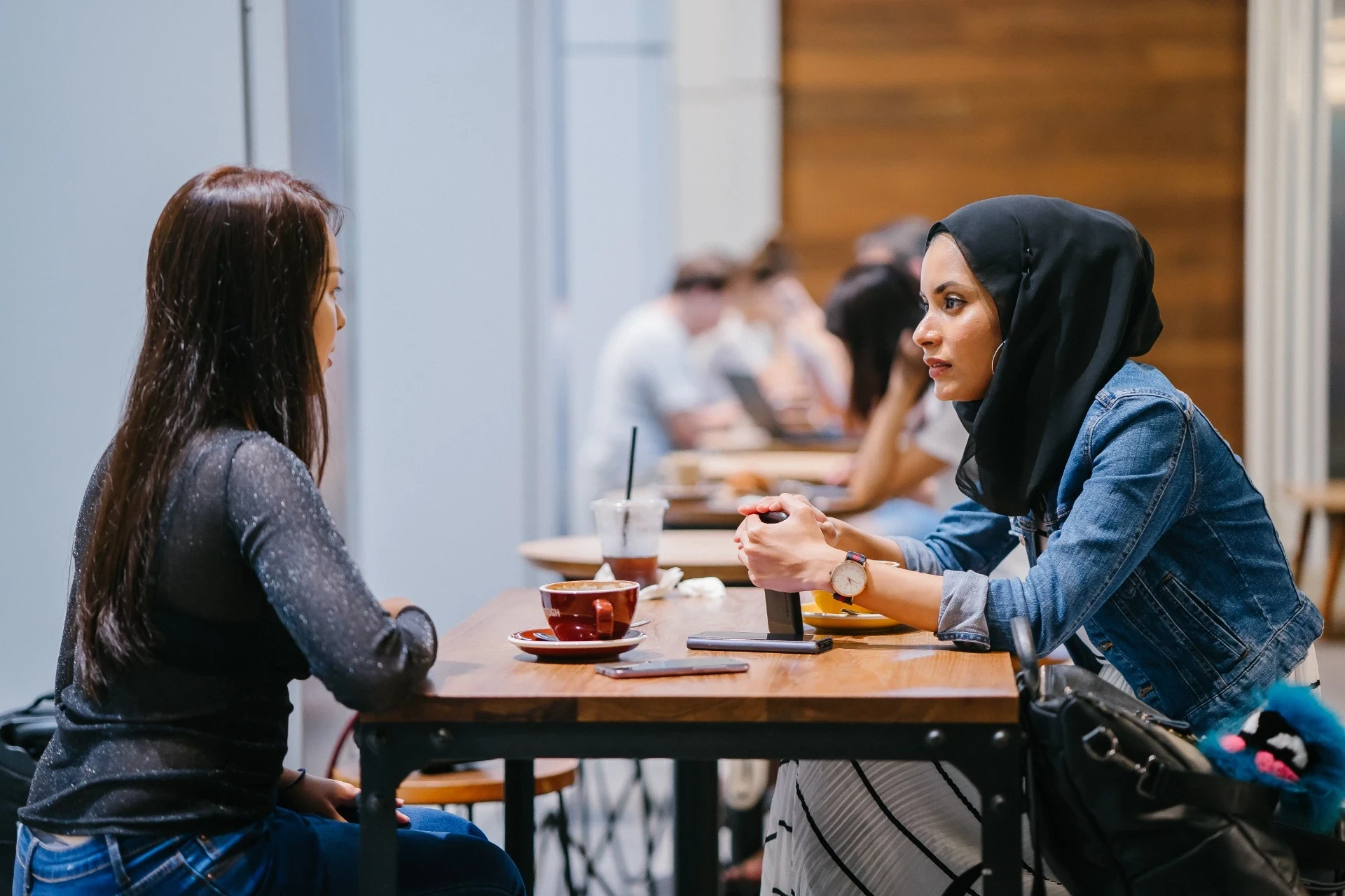 Imagem de duas mulheres sentadas frente a frente em uma mesa de café, envolvidas em uma conversa séria. Uma delas, usando hijab e jaqueta jeans, está com as mãos cruzadas e parece focada na conversa, enquanto a outra está com uma expressão atenta. Sobre a mesa, há xícaras e copos de café, e no fundo é possível ver outras pessoas em um ambiente descontraído, provavelmente um café ou espaço de trabalho colaborativo.