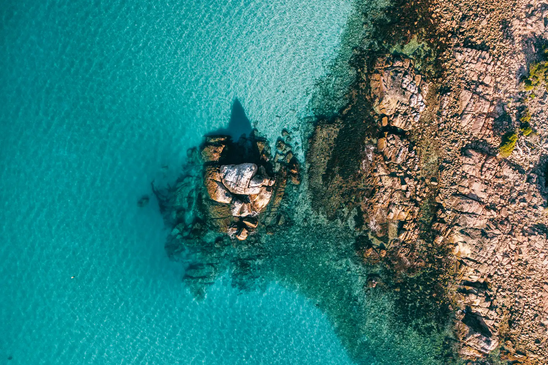 Aerial of a granite outcrop in clear turquoise water beside a rocky shore