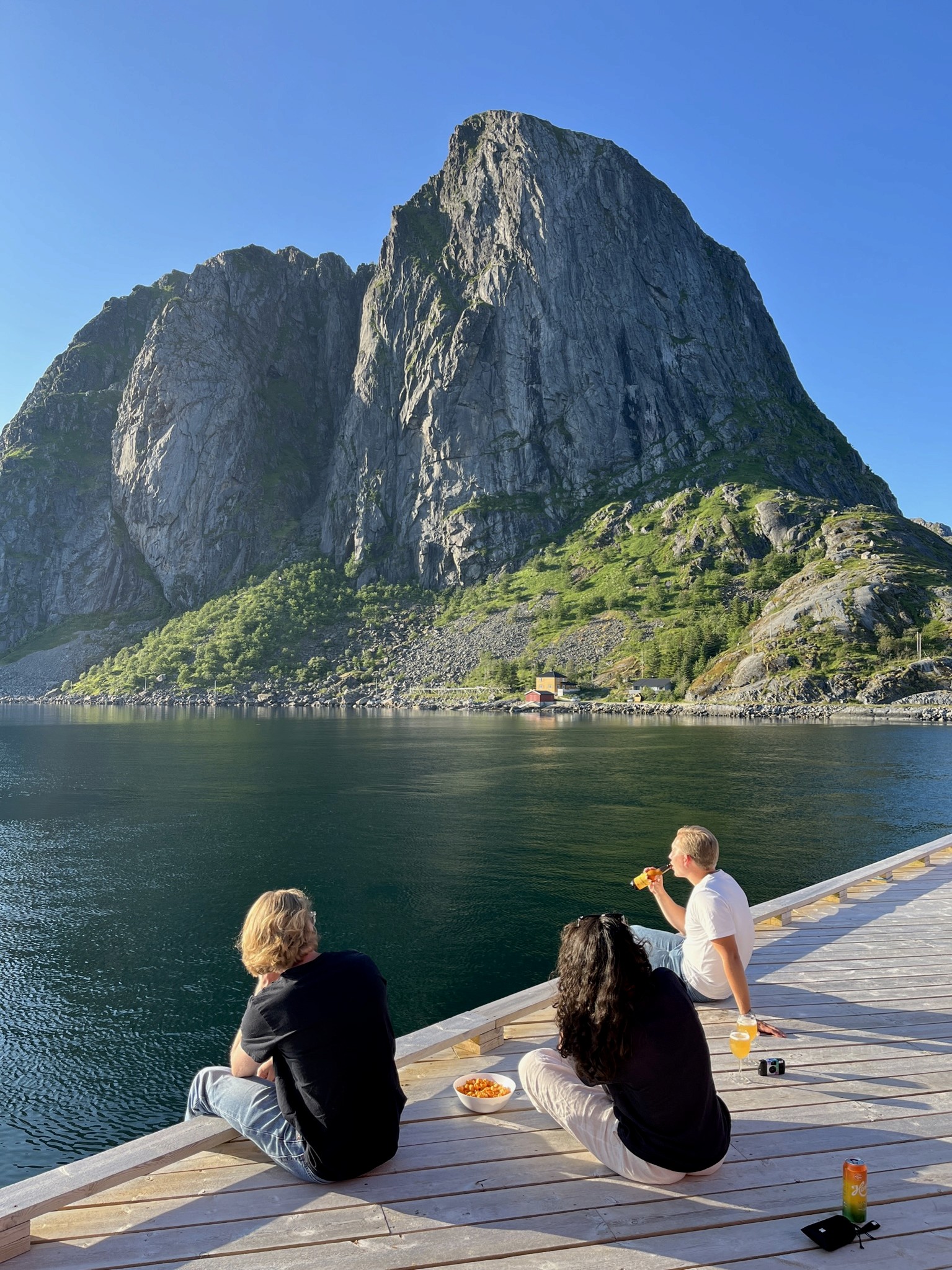 3 people sitting on a jetty with a view over water and mountains