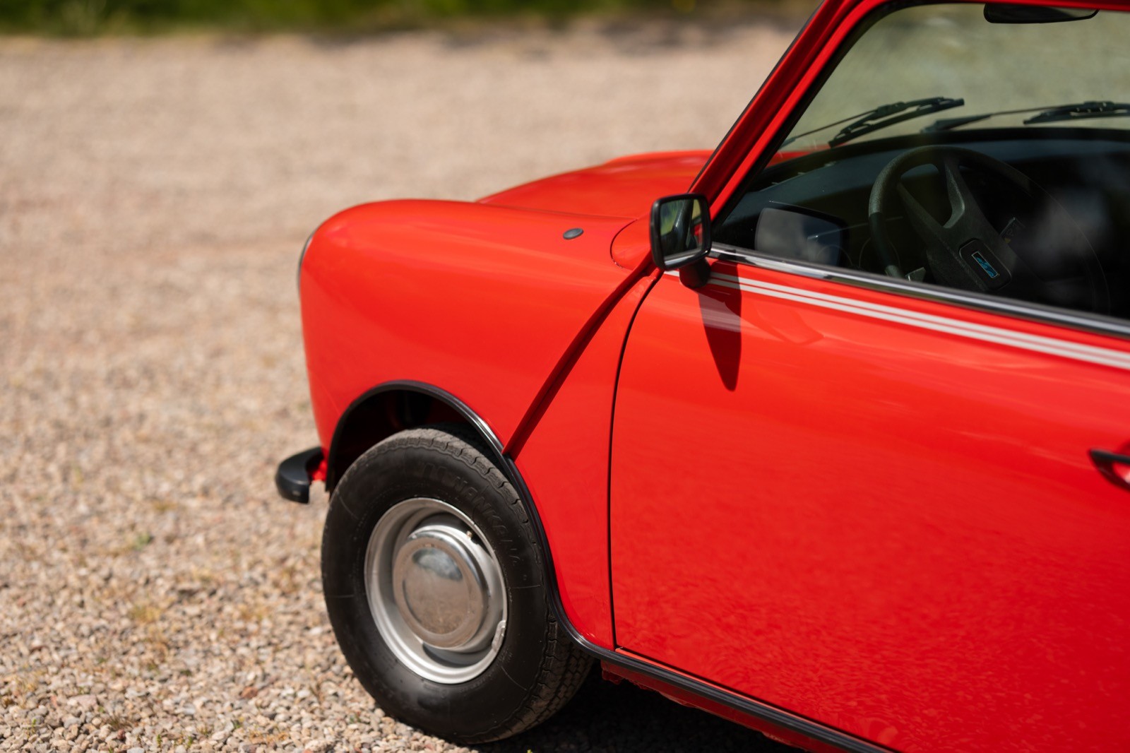 Close-up of a bright red vintage car, showcasing the front wheel and door, parked on a gravel surface.