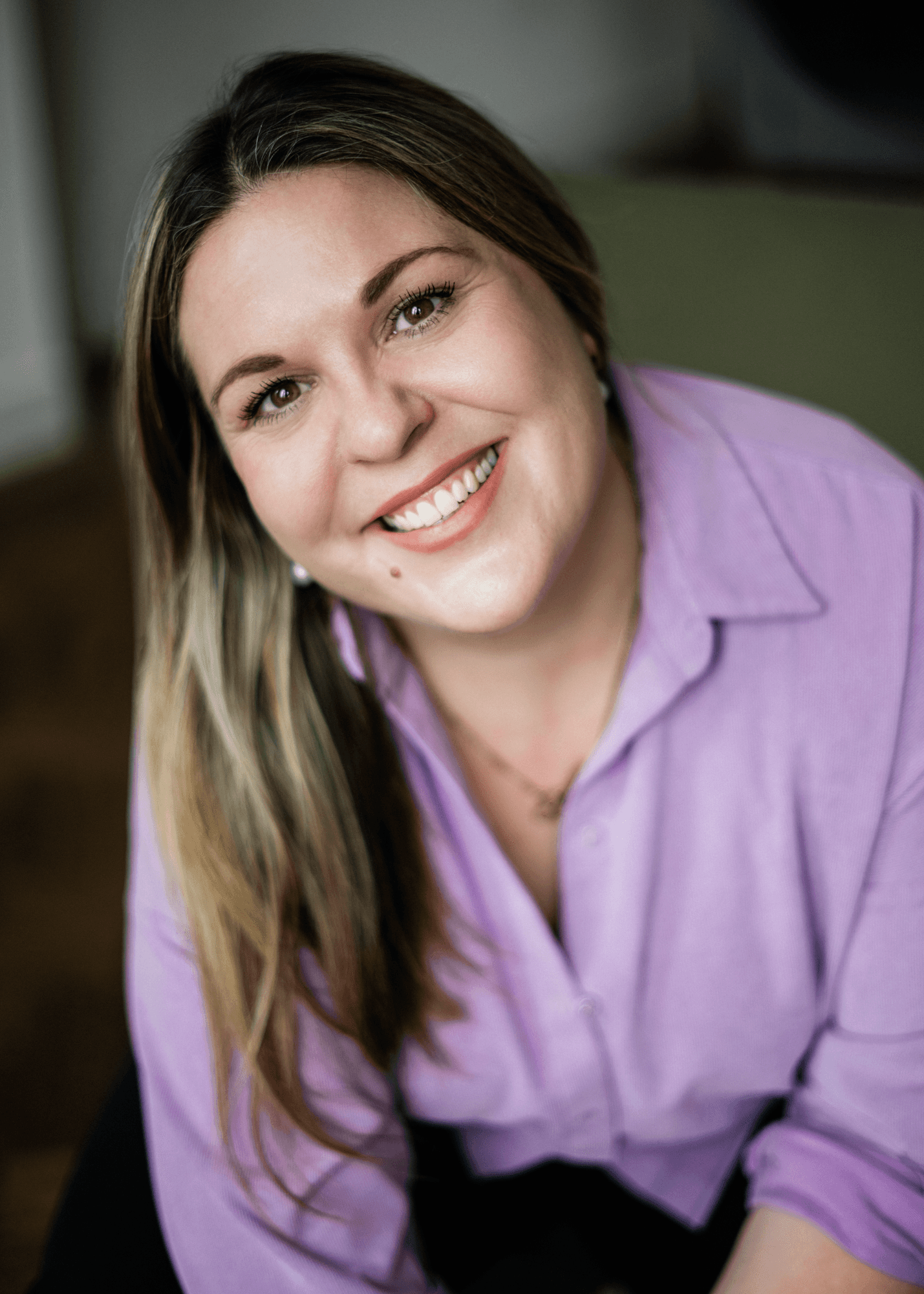 headshot casting photo of Louise O’Meara, actor, smiling woman with long brown hair, lilac blouse, dark background.