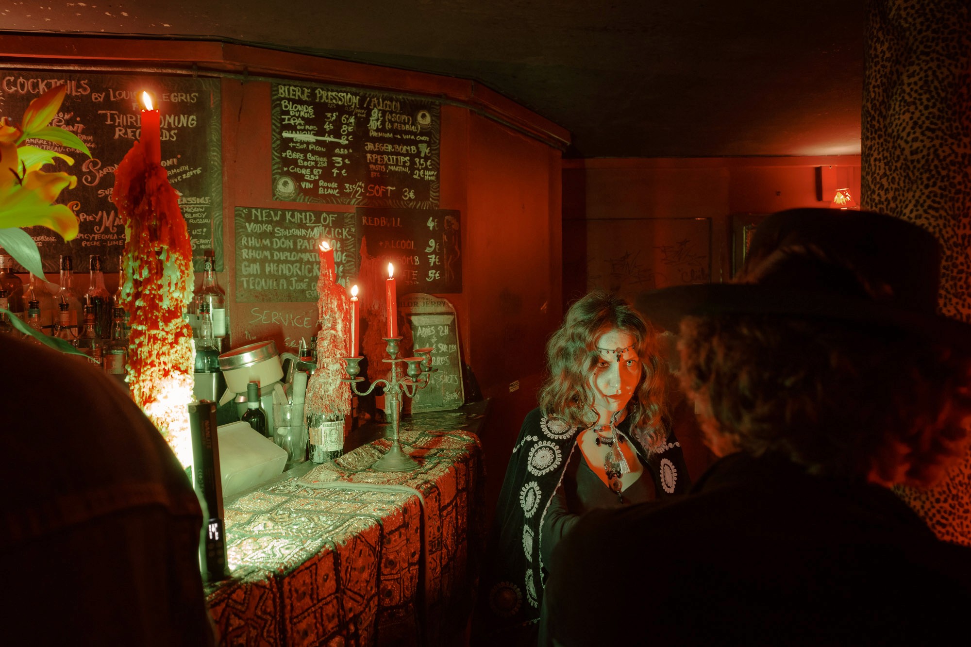 A dimly lit bar with a warm, vintage ambiance features a rustic counter adorned with intricate patterns, red candles flickering atop, and a detailed cocktail menu on a wooden wall, creating a mysterious, cozy atmosphere.