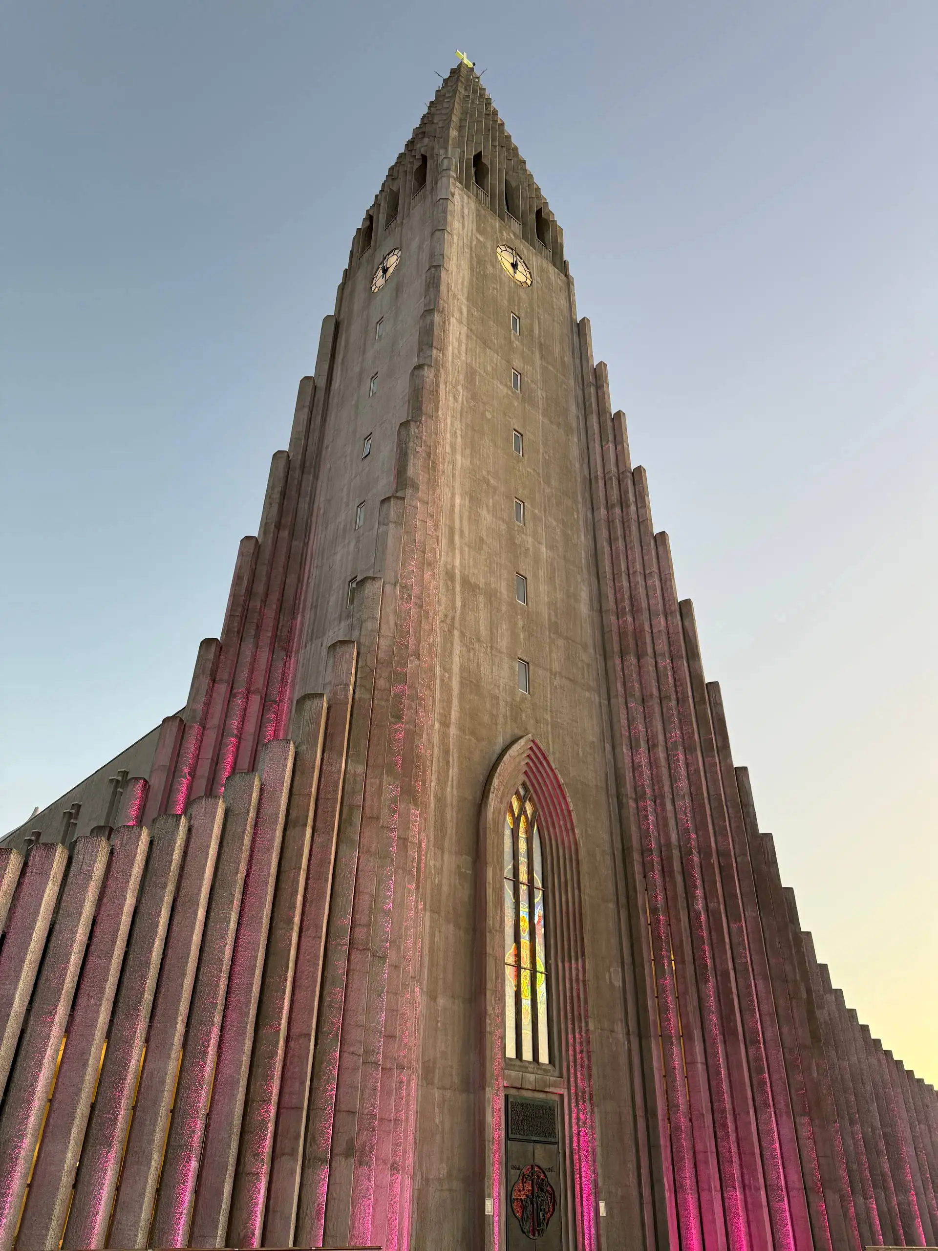 Hallgrímskirkja church in Reykjavík seen from below, its stepped concrete façade lit pink at sunset beneath a clear blue sky