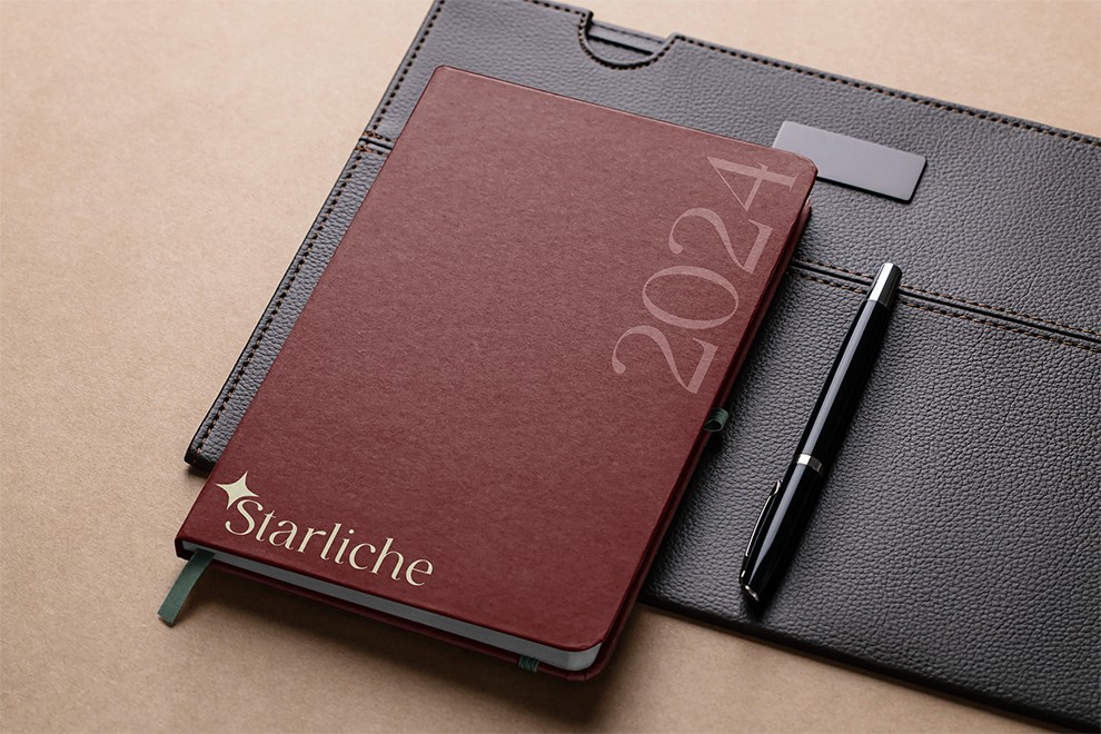 Premium corporate stationery design mockup featuring a bespoke dark red leather notebook embossed with the Starliche brand logo and the year 2024 resting next to a sleek executive pen.