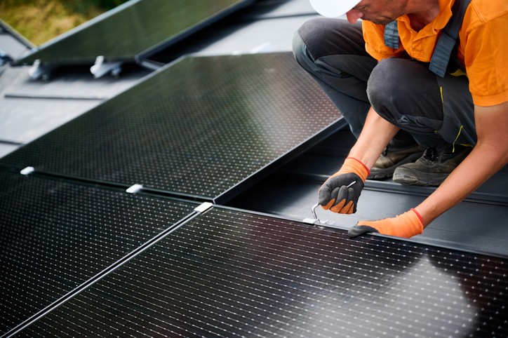 Eine Person installiert Solarpaneele auf einem Dach und hantiert konzentriert mit einem schwarzen Modul.