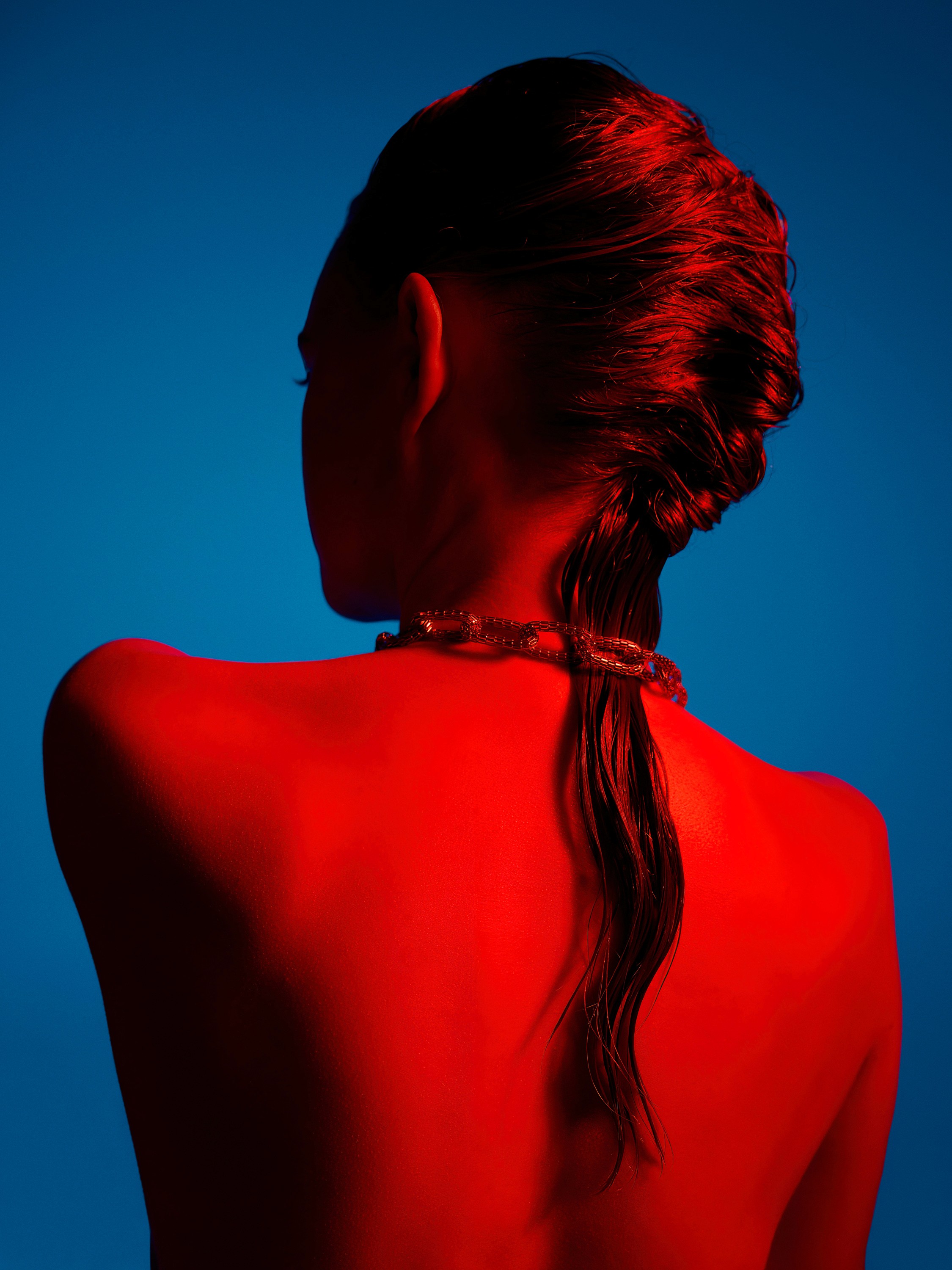 The back of a female model with a braid and heavy chain necklance bathed in red light on a blue background.