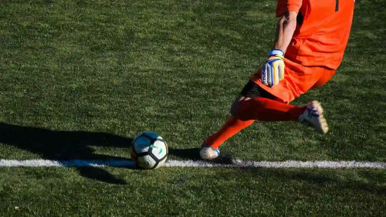 A modern goalkeeper in bright orange executes a pass during a game, demonstrating the evolutionary role of the goalkeeper as part of the build-up play.