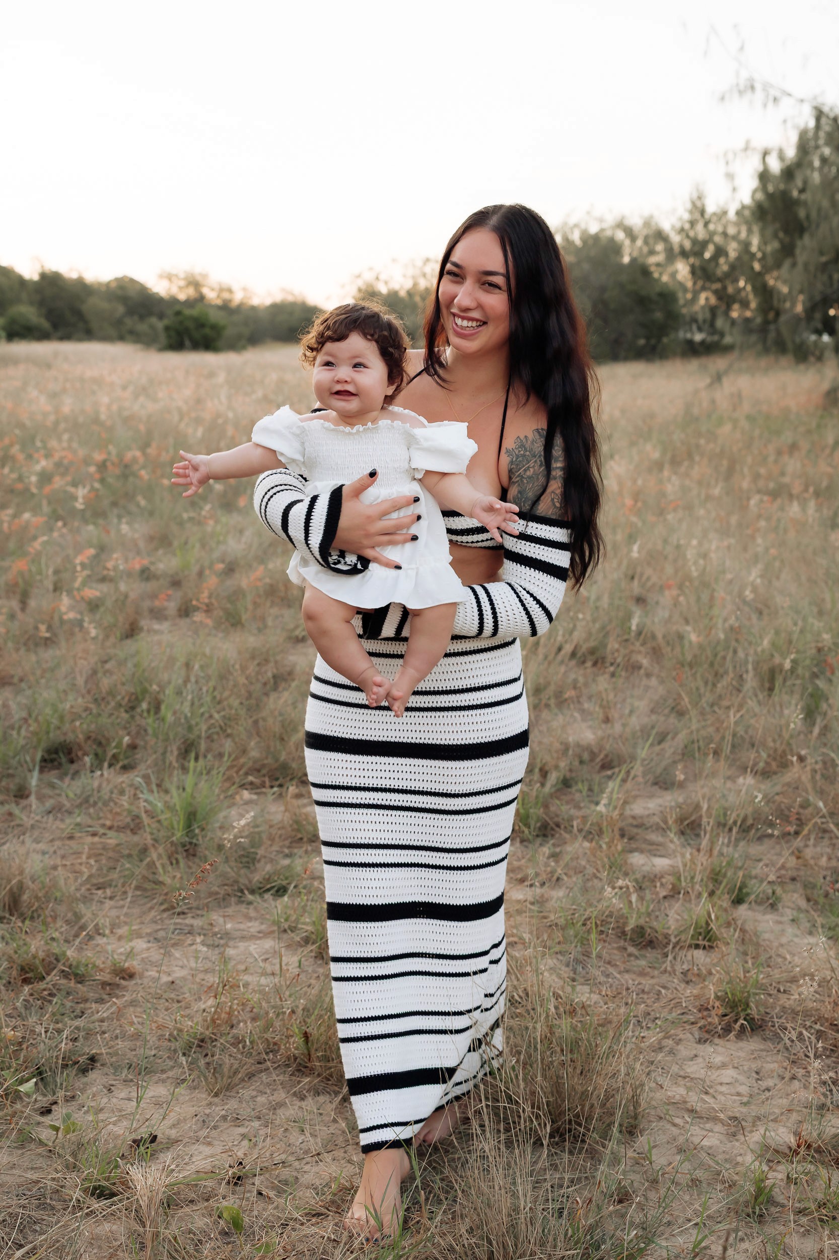 Mother and child standing together in tall grass at sunset, photographed during a soft outdoor motherhood session in Mackay.