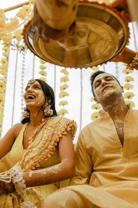 Spontaneous shot of a couple laughing during their haldi ceremony, with faces adorned in turmeric paste, showcasing the fun and cultural essence in candid wedding photography.