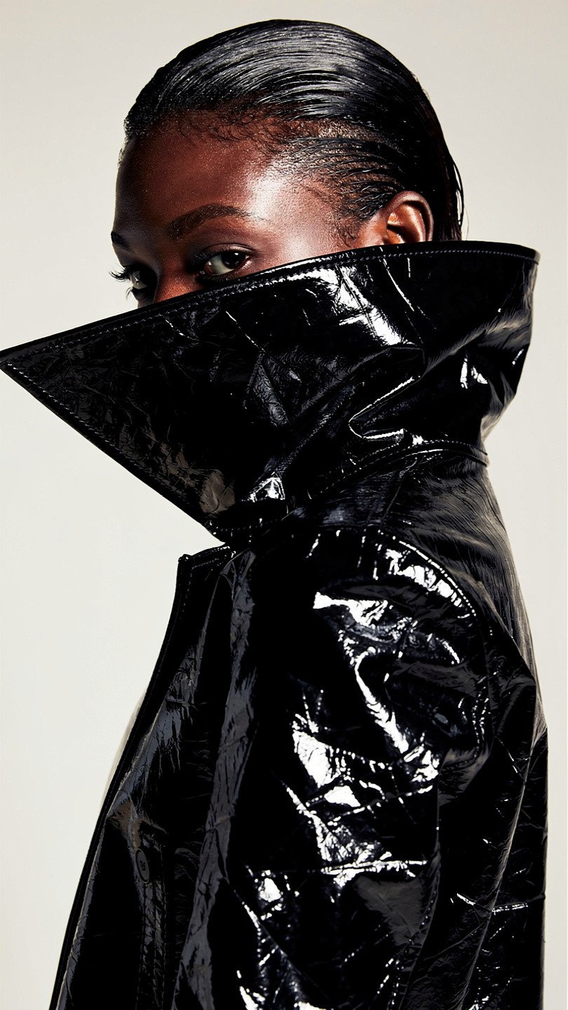 A person with slicked-back hair wears a glossy black trench coat with a high collar, looking serious and intense against a neutral background.