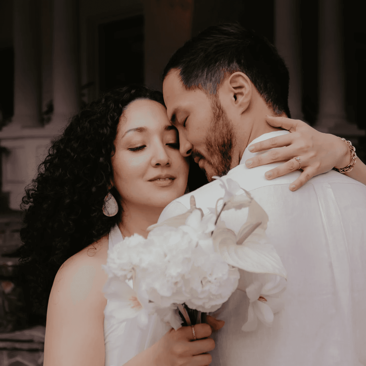 A bride and groom hold hands tenderly, the bride's bouquet of dried flowers hanging low. They stand in a softly lit, elegant interior, conveying romance.