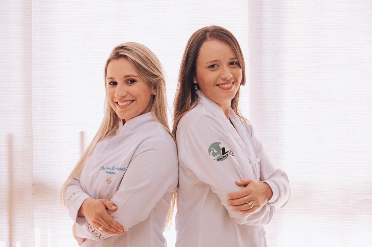 Two doctors in white lab coats and stethoscopes standing confidently in a clinical setting with medical posters and a bed in the background