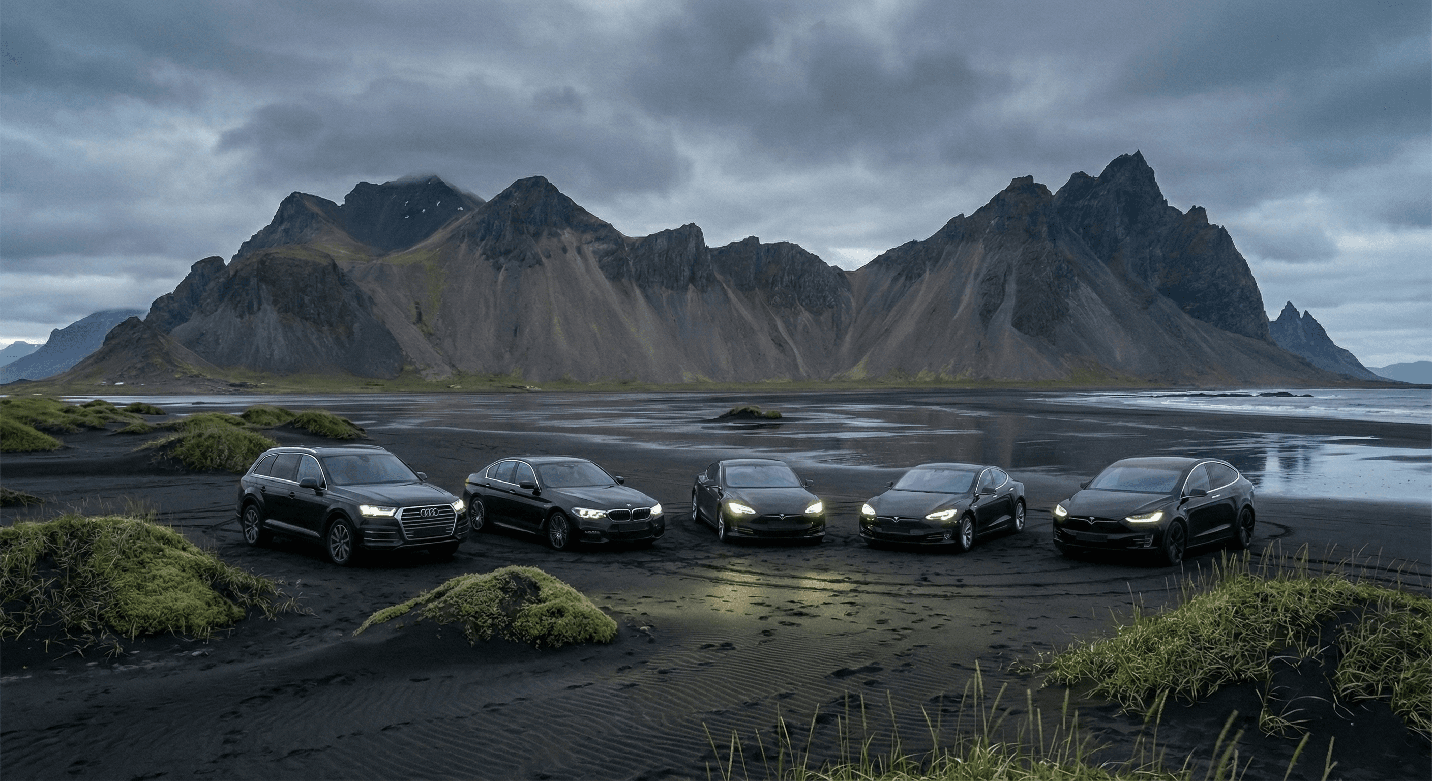 A fleet of five dark luxury cars, including Audis and Teslas, parked in a row on a black sand beach with jagged mountains in the background.