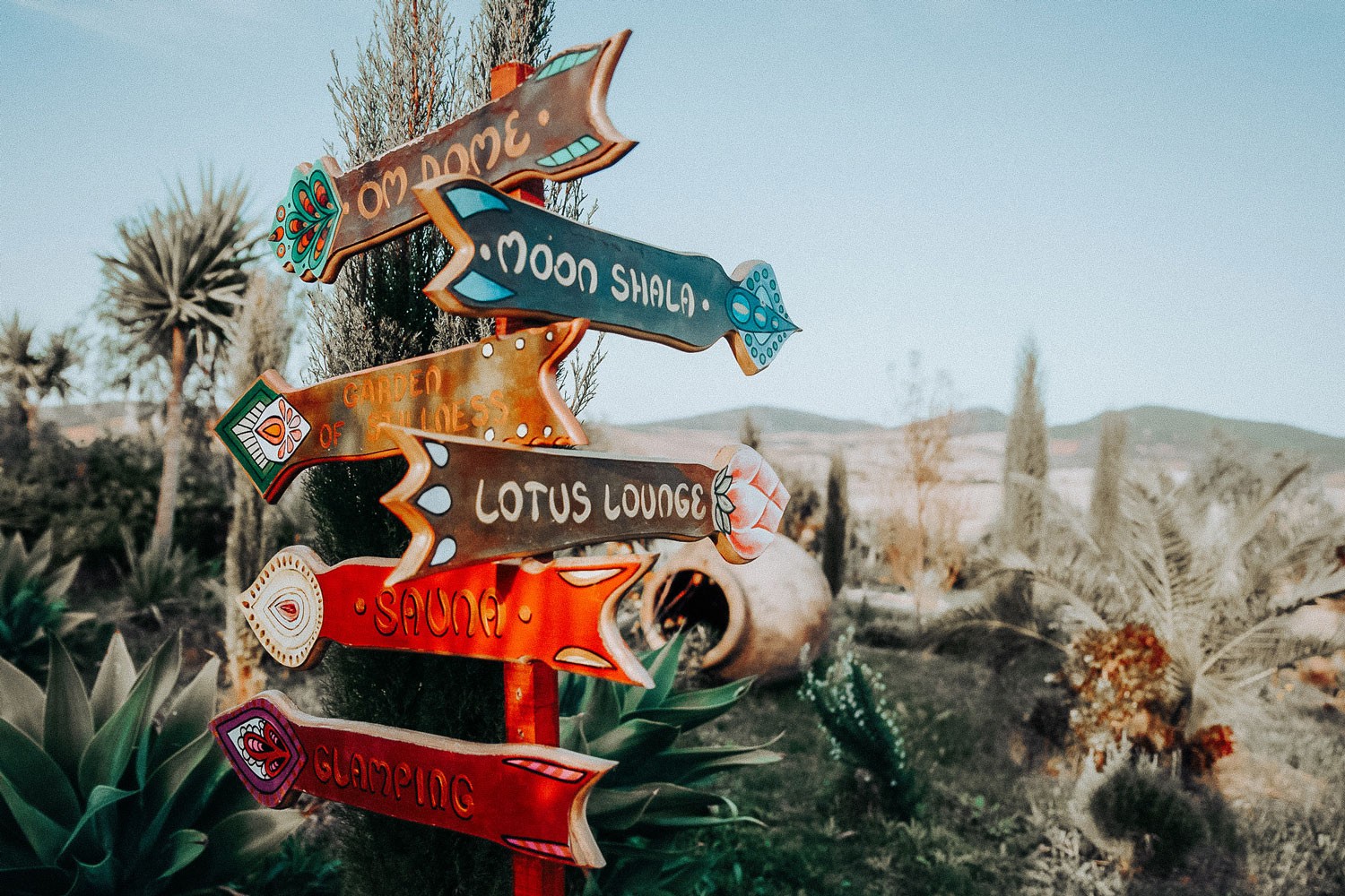 Colorful wooden signpost with arrows to "Moon Shala," "Lotus Lounge," and more, set in a desert-like garden environment. A relaxed, whimsical mood.
