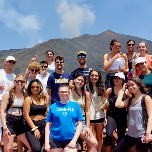 Um grupo de 16 pessoas posando em frente a um vulcão fumegante sob um céu azul claro.