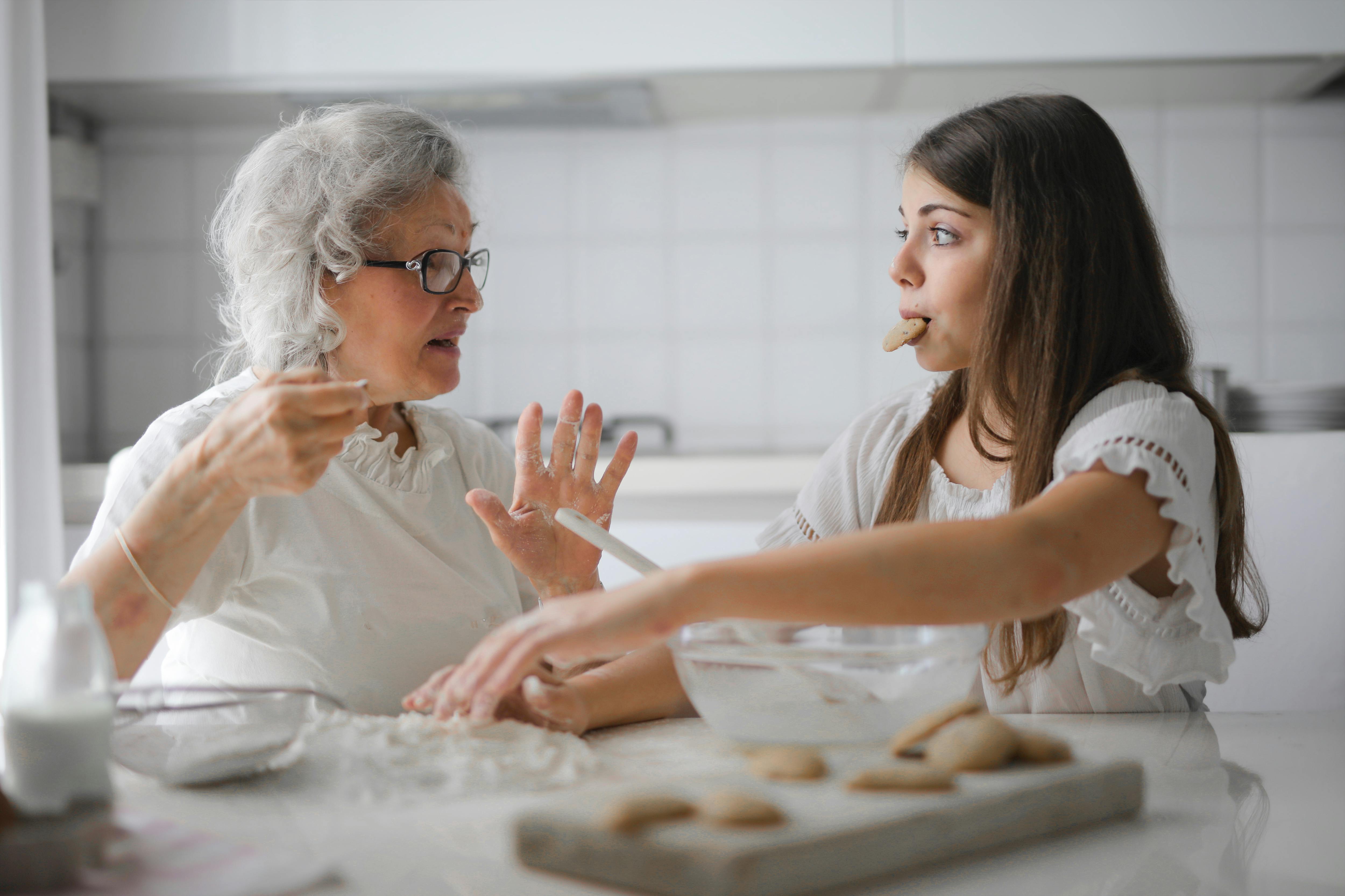 A younger woman supporting an older woman with gentle conversation and hand gestures in a kitchen with baked goods.