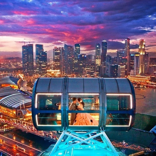 A couple in formal attire sits inside a glass observation pod during sunset, overlooking a cityscape with illuminated skyscrapers.