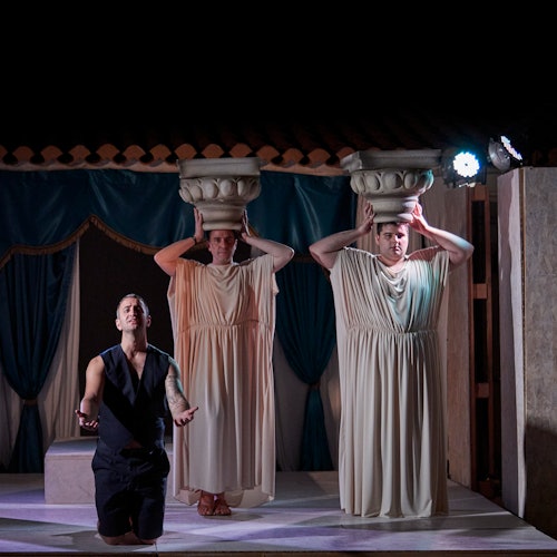 舞台上有三位演员;其中一人跪着,双臂张开,另外两人站着,头上顶着柱头。