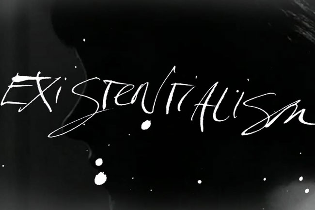 The image features the phrase "EXISTENTIALISM" written in white cursive font against a black background. The text is centrally positioned and spans across the width of the image. White splatters or paint-like marks are scattered around the text, adding visual texture. The background is predominantly black, with subtle variations in shade visible around the text and splatters.