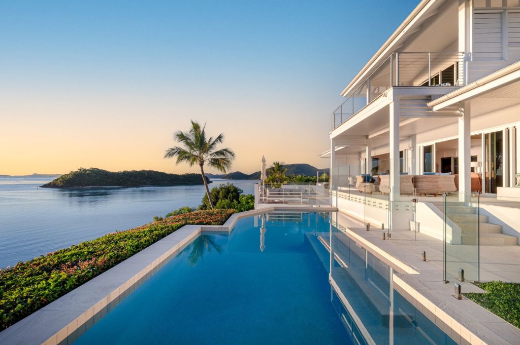 Luxurious oceanfront villa with a modern design, featuring an infinity pool overlooking a tranquil sea at sunset, surrounded by lush greenery and distant islands.