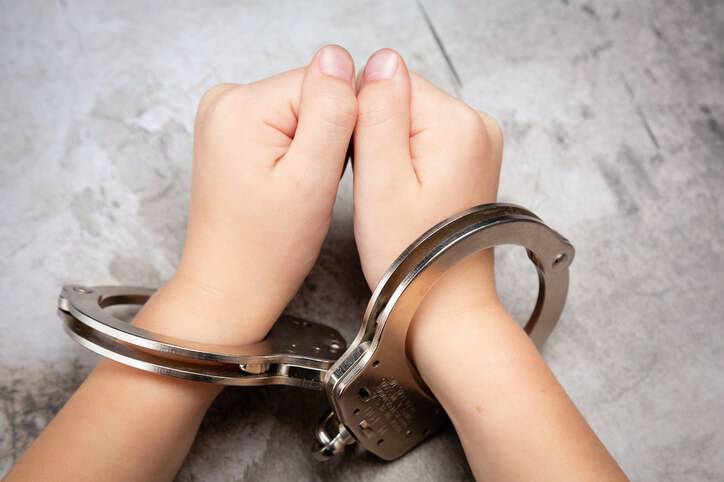 Two juvenile hands are clasped together, secured in a pair of metal handcuffs against a textured gray background, reflecting legal themes of arrest and detention.