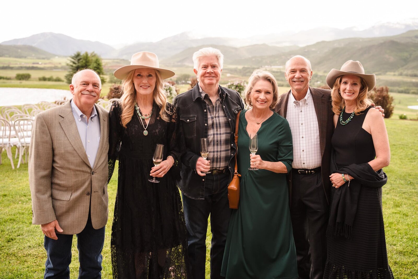 Six guests posing on a lawn with ceremony chairs, water, and mountains behind them
