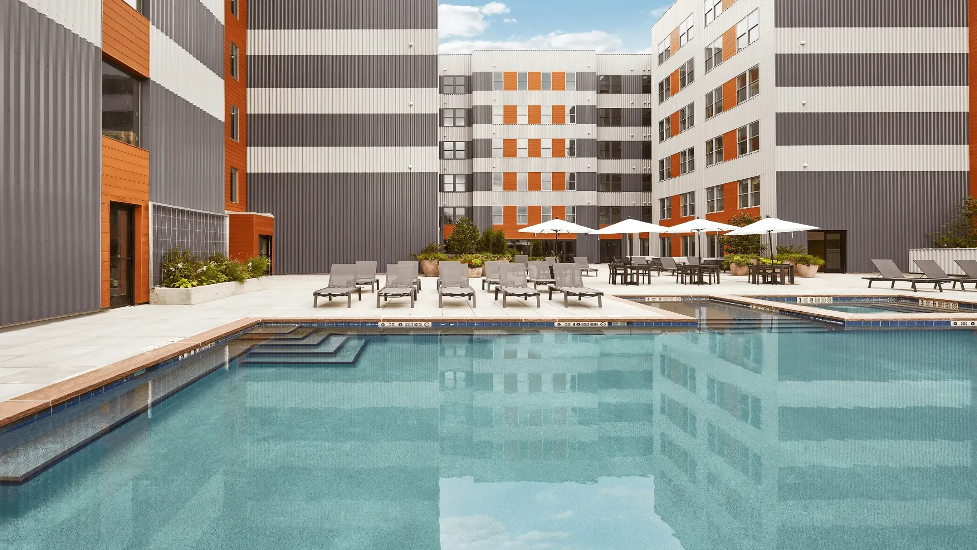 Modern apartment complex with a swimming pool and lounge chairs under blue skies, surrounded by buildings.