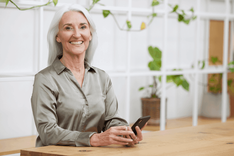 Smiling elderly woman with long gray hair holding a smartphone while seated at a table in a light, modern room.