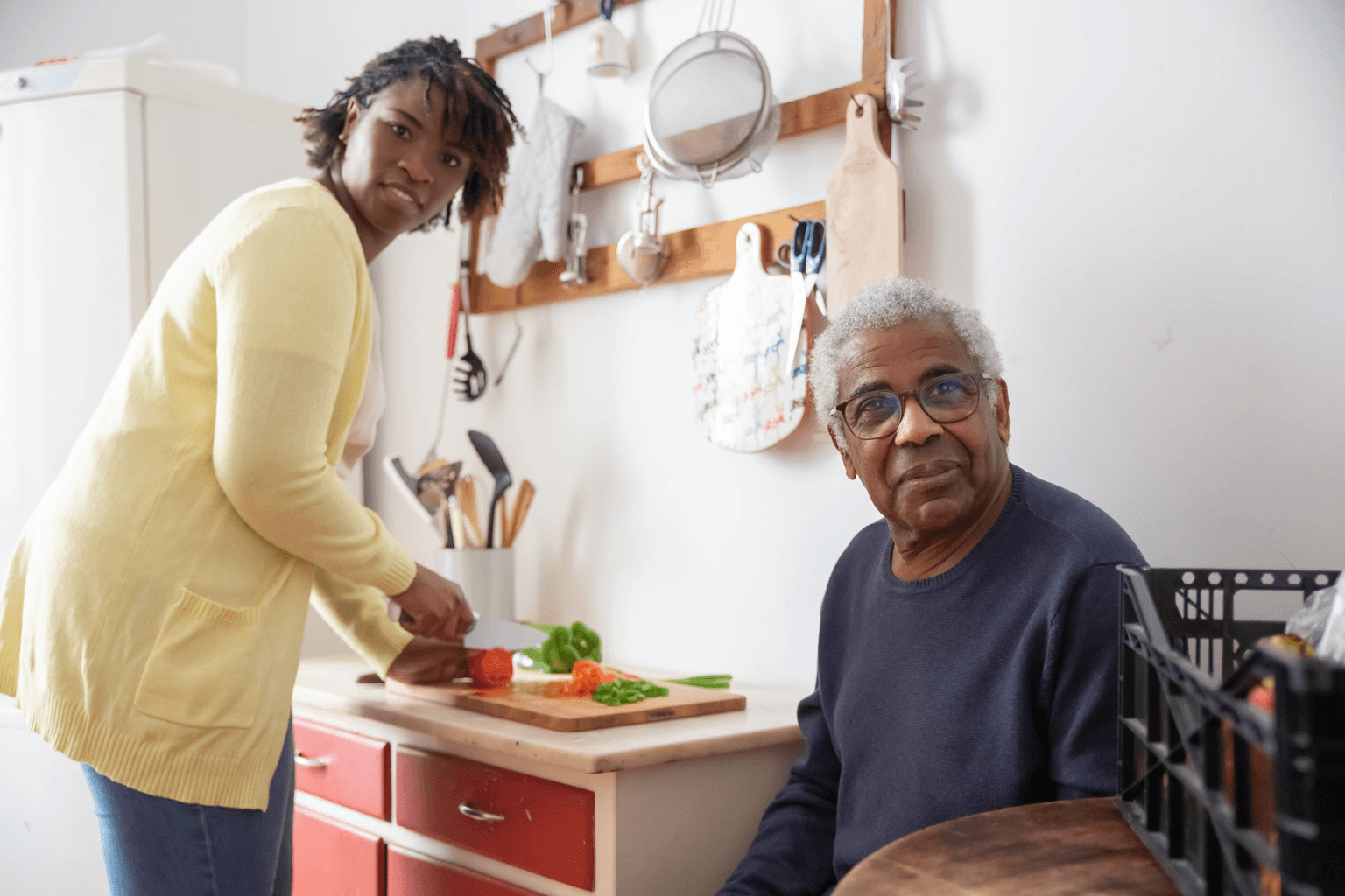 A home care aide chopping vegetables at the counter while the older man she visits sits at his kitchen table.