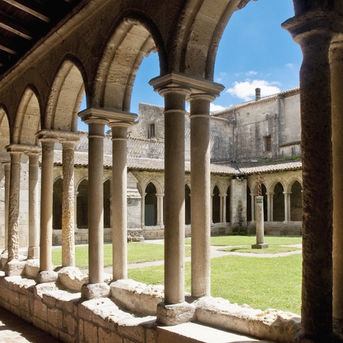 Stone arches frame a courtyard with grass and a statue, surrounded by historic buildings under a clear blue sky.