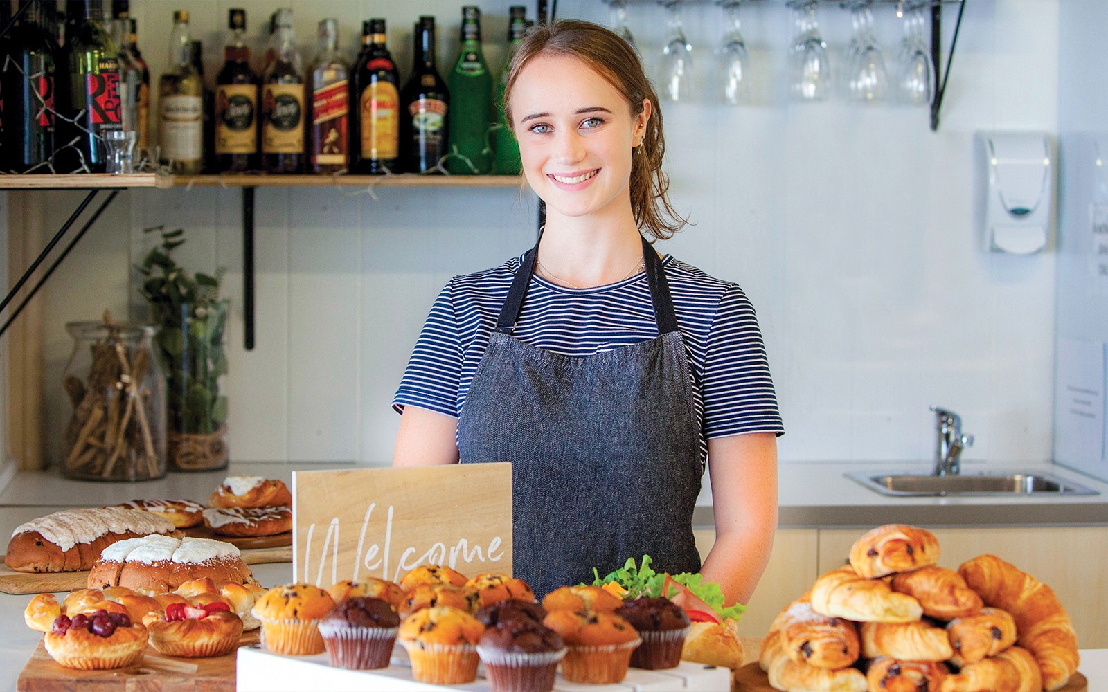 Bakery display with pastries and muffins on a counter, staff member smiling behind.