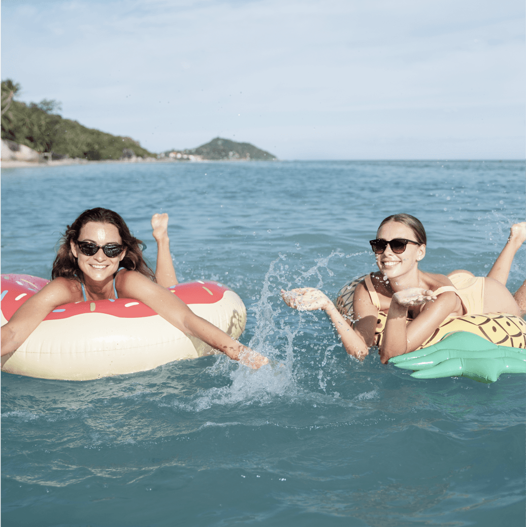 Two women relaxing on inflatable floats and splashing water in a sunny ocean.