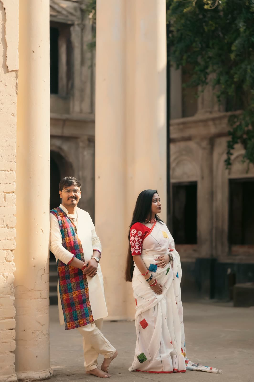Couple standing apart between stone columns in white and patterned clothes