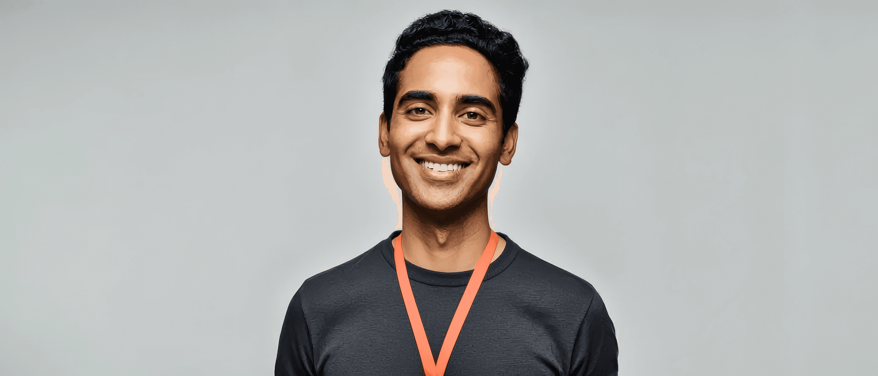 A man wearing a dark shirt and an orange medal ribbon stands in front of a plain light background, smiling at the camera.