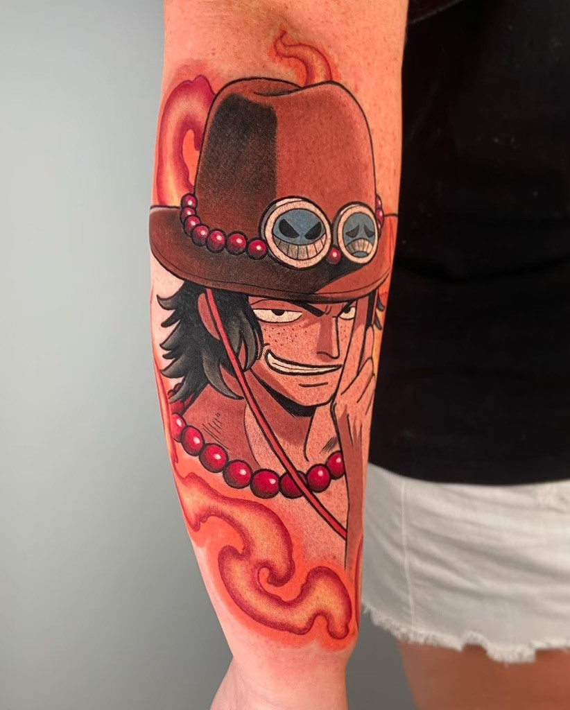 A vibrant tattoo of Portgas D. Ace from One Piece, showcasing his iconic hat adorned with goggles and smiling skull symbols. The fiery effects and beads around his neck highlight his flame-based abilities, while the tattoo’s placement on the forearm adds dynamic visibility.