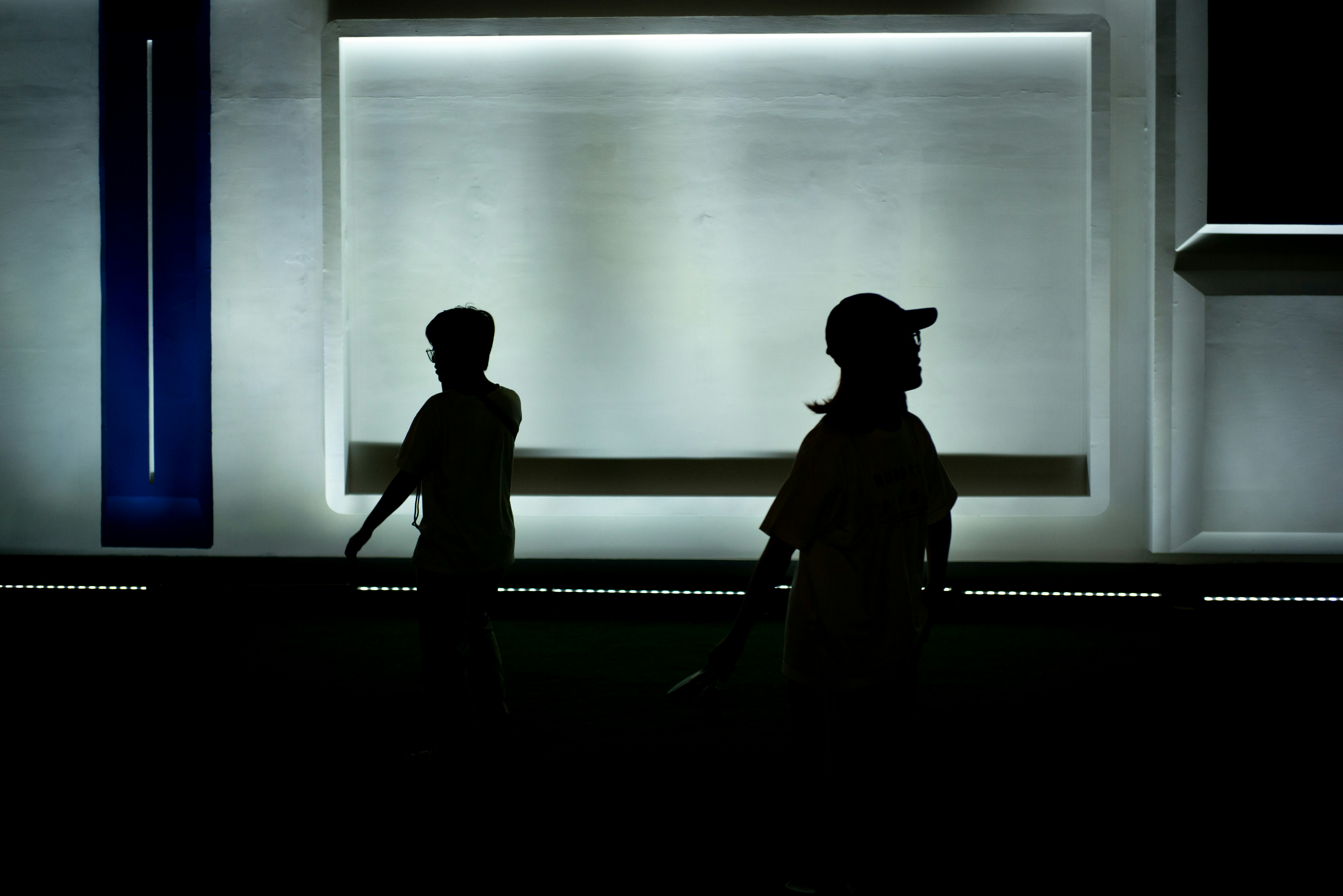 A silhouette of two people walking in front of a large, glowing rectangular light installation on a dark wall.