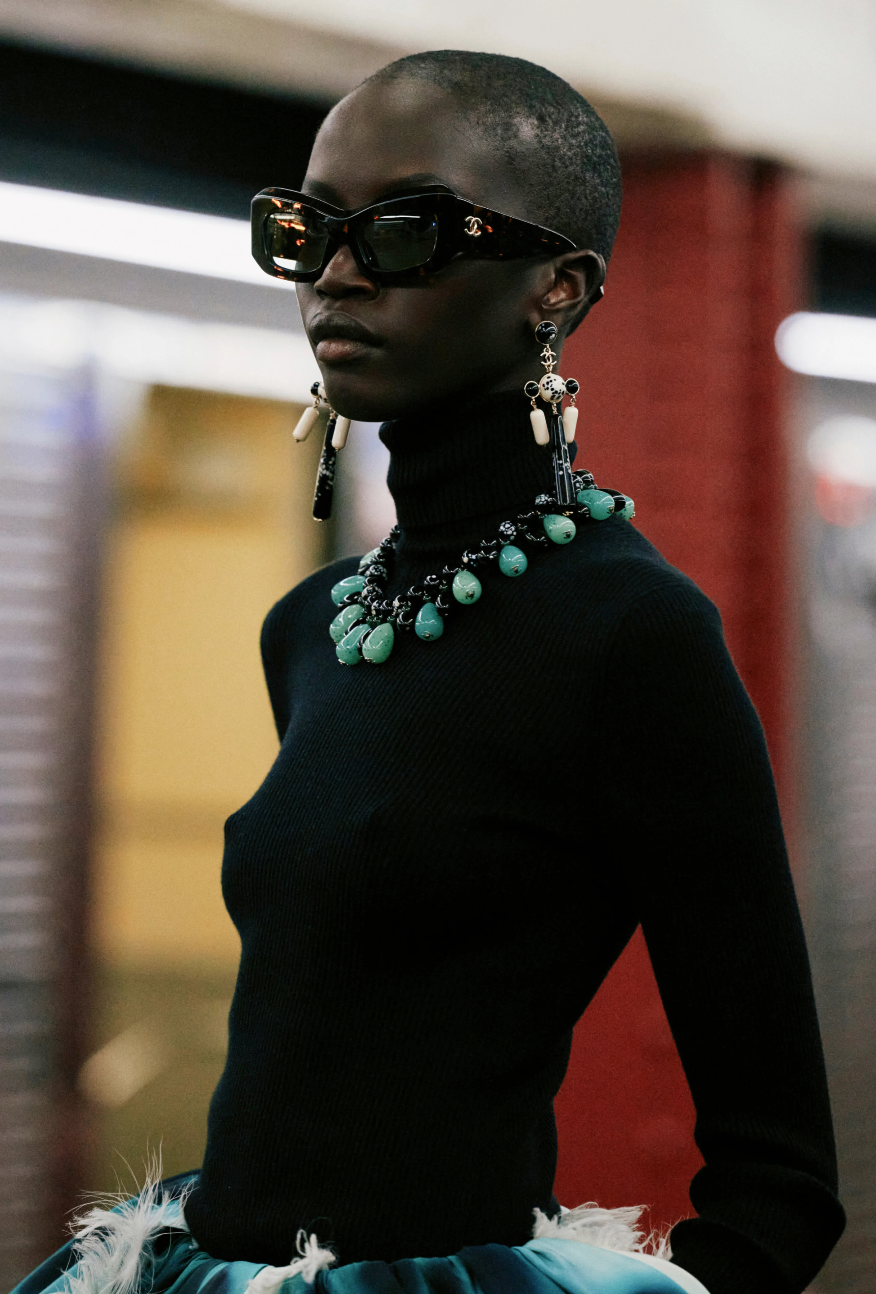 A model with a shaved head wears dark sunglasses, a black high-neck outfit, and statement turquoise jewelry at the Chanel Métiers d'Art 2026 show.