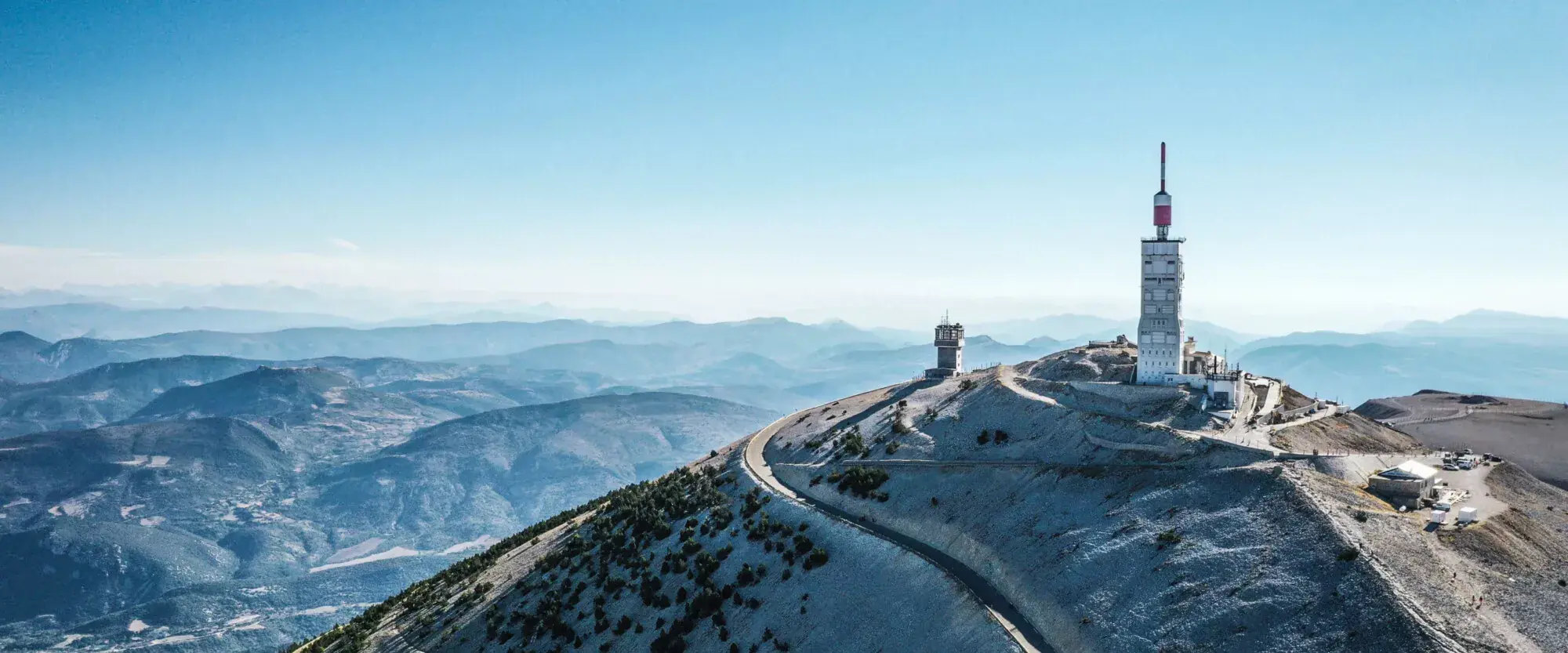 Vue aérienne panoramique du sommet du Mont Ventoux sous un ciel bleu clair, montrant l'observatoire et son antenne de télécommunication rouge et blanche dominant un paysage de crêtes calcaires arides et de montagnes lointaines.