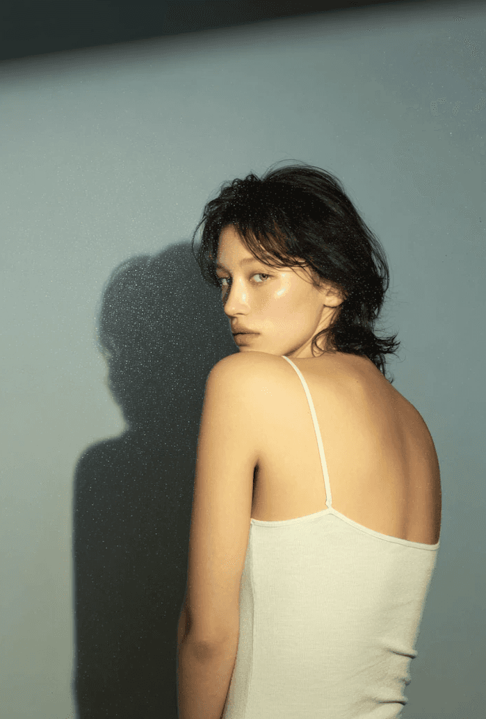 Portrait of a woman in a white tank top looking over her shoulder with dramatic lighting and shadow on the wall.