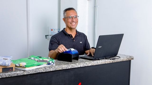 Homem de óculos e cabelos grisalhos, utilizando notebook e impressora de cupom fiscal sobre balcão de loja com roupas dobradas.